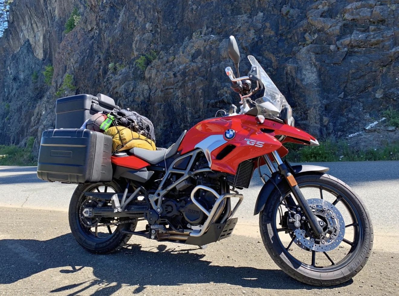 A red BMW F 700 GS adventure motorcycle with panniers and a loaded tail pack, parked beside a rock cliff on a coastal road