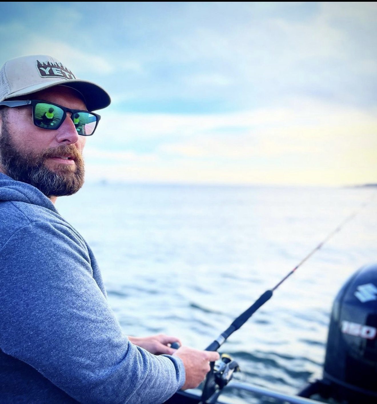 A man in sunglasses fishing from a boat on calm water at dawn