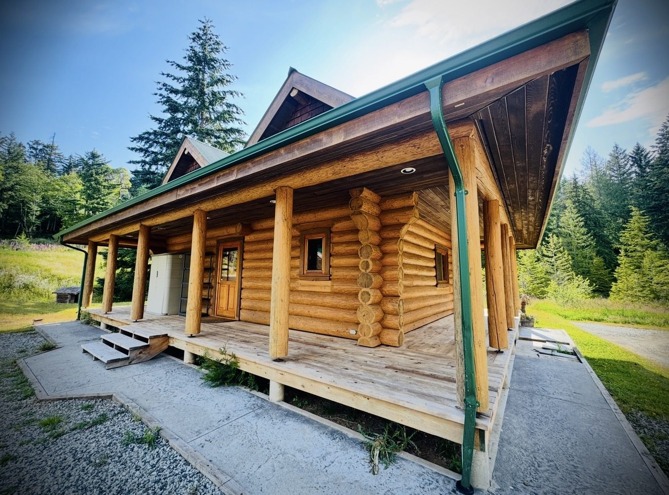 A log cabin with a wide covered porch, green roof and forest behind