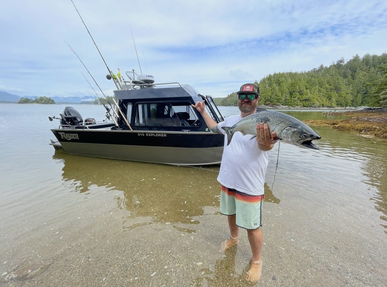 A man holding up a freshly caught salmon in front of a fishing boat on a calm, shallow bay