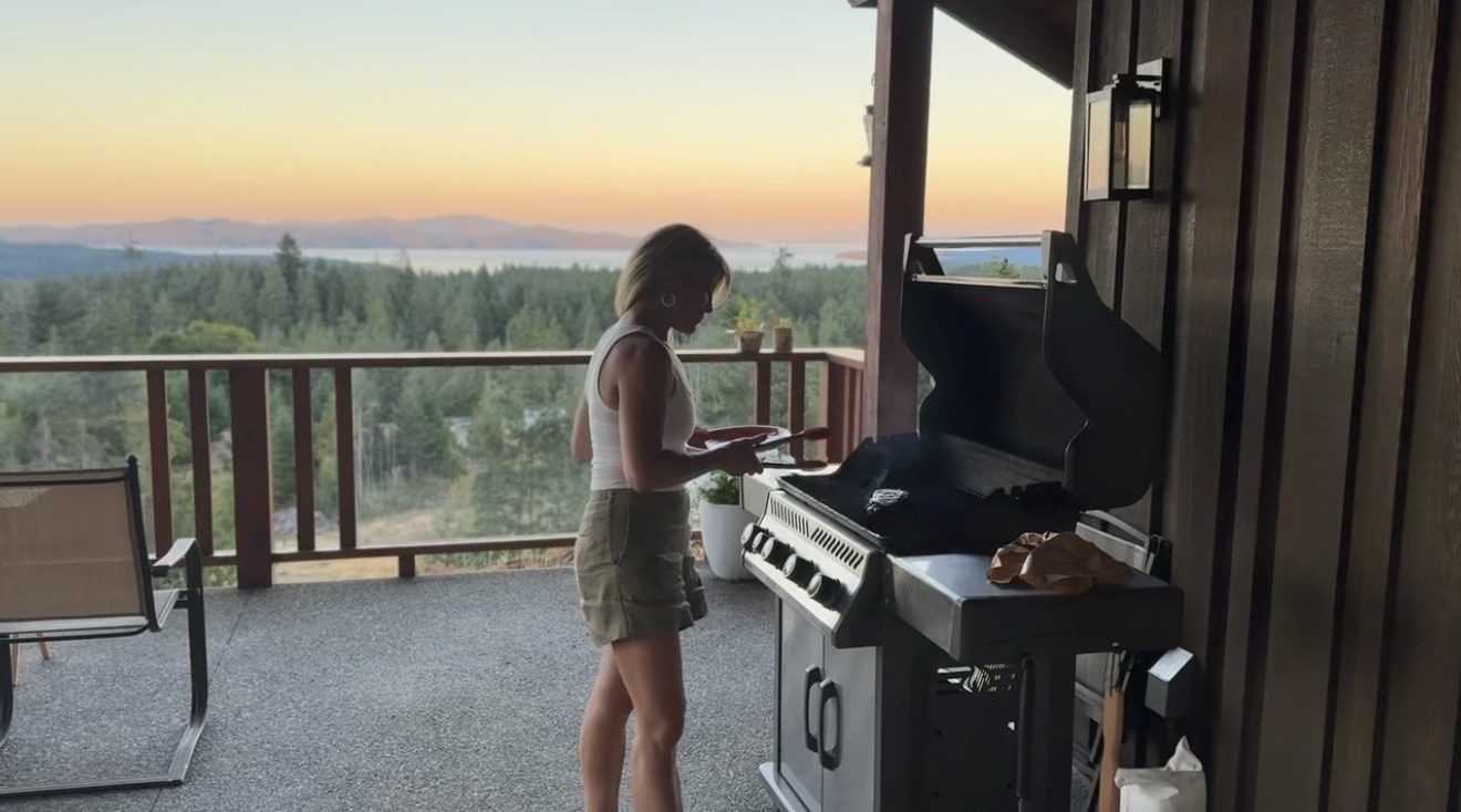 A covered deck with a barbecue overlooking forest and sea at sunset