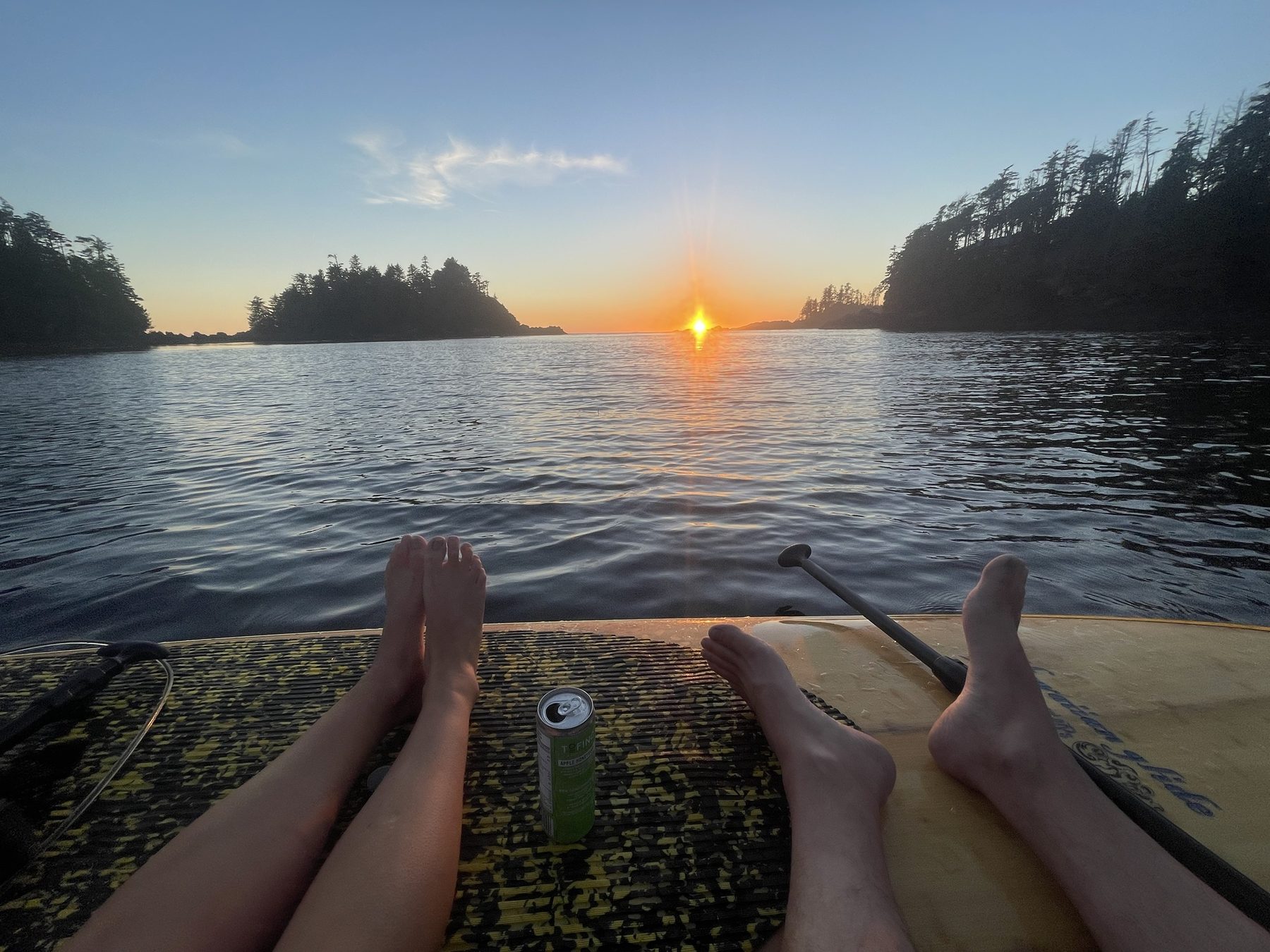 Two pairs of feet resting on paddleboards facing a sunset between forested headlands
