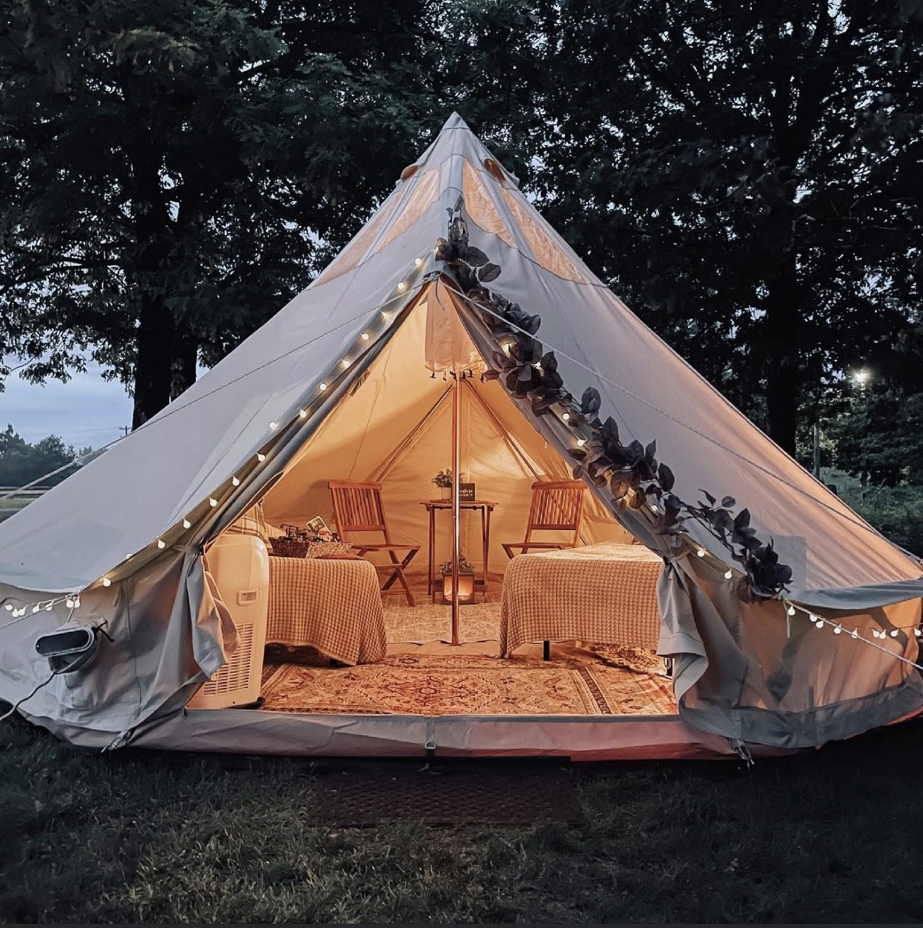 A canvas wall tent glowing at dusk, with string lights and two beds, rugs and chairs inside